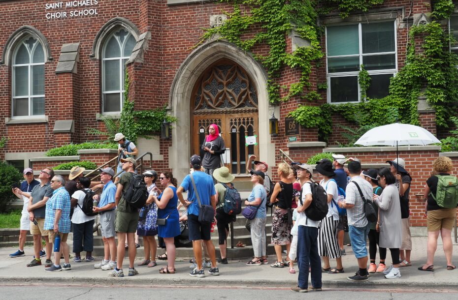 A large group of people in summer attire are standing along a sidewalk looking across road and are in front of a red brick building that features windows with pointed arches.