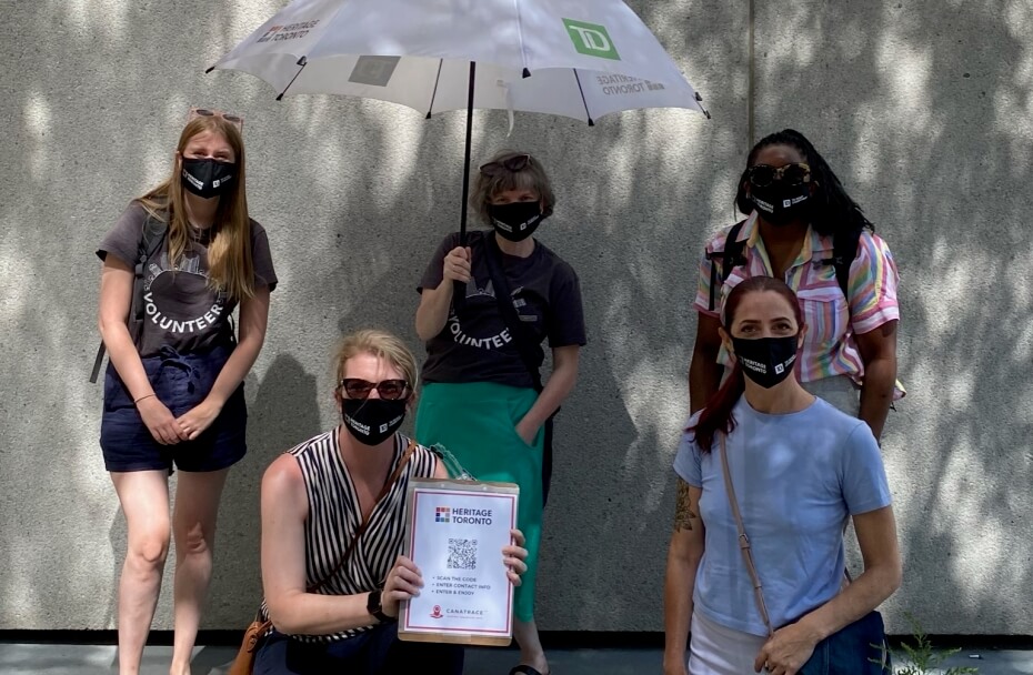 Five women stand and crouch facing the camera against a grey concrete building. They all are wearing masks. The woman in the centre back holds a large white umbrella.