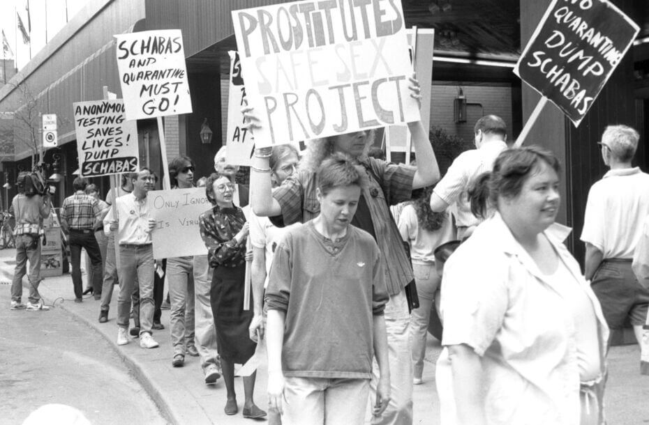 A black-and-white photograph of a group of protestors. They are all walking in a growp along a path. Six of them are carrying signs protesting protesting Ontario Medical Officer of Health Richard Shabas and quarantines.