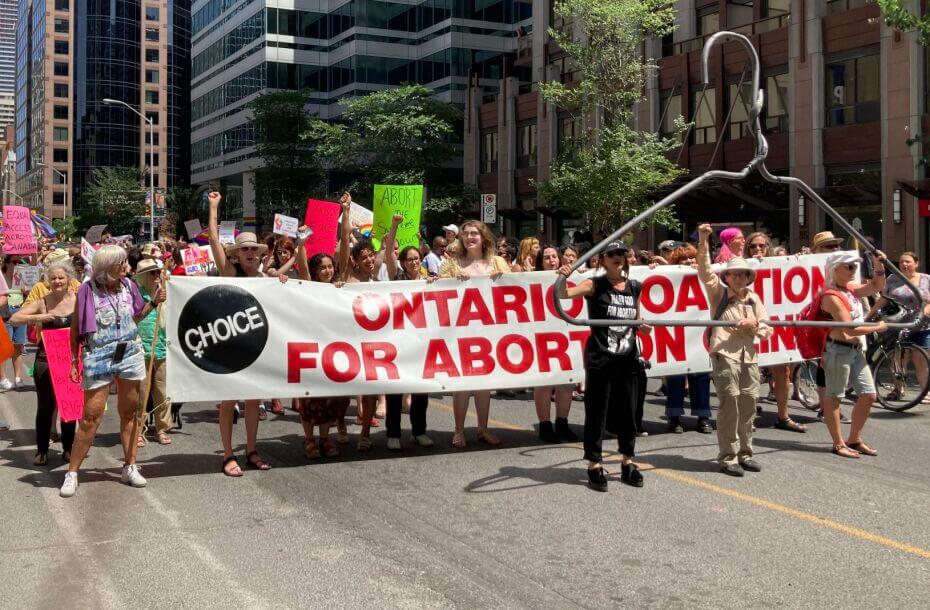 A large group of people, mostly women, walk down a city street. At the front of the group, individuals carry a large white banner with red lettering that reads 