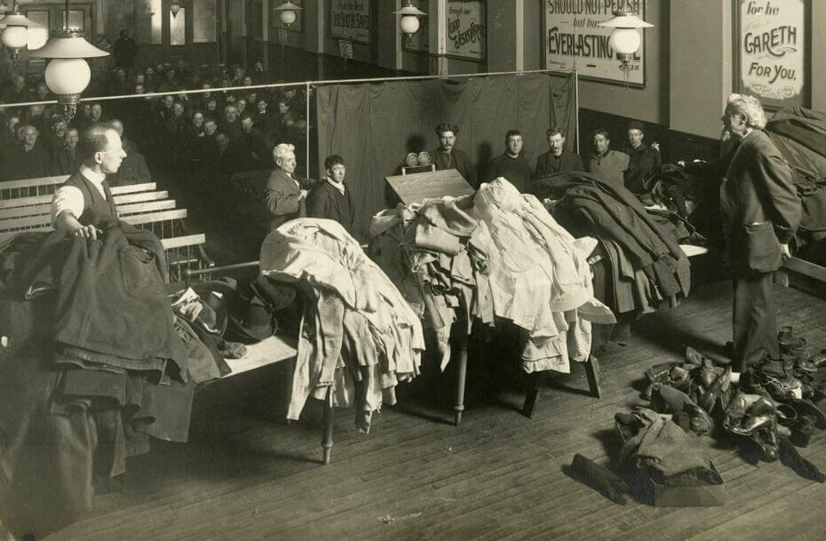 A group of men sitting in a room. In the front is a line of men waiting to go up to an area with clothes. More men are standing with with tables full of clothing (shirts, pants, and shoes).