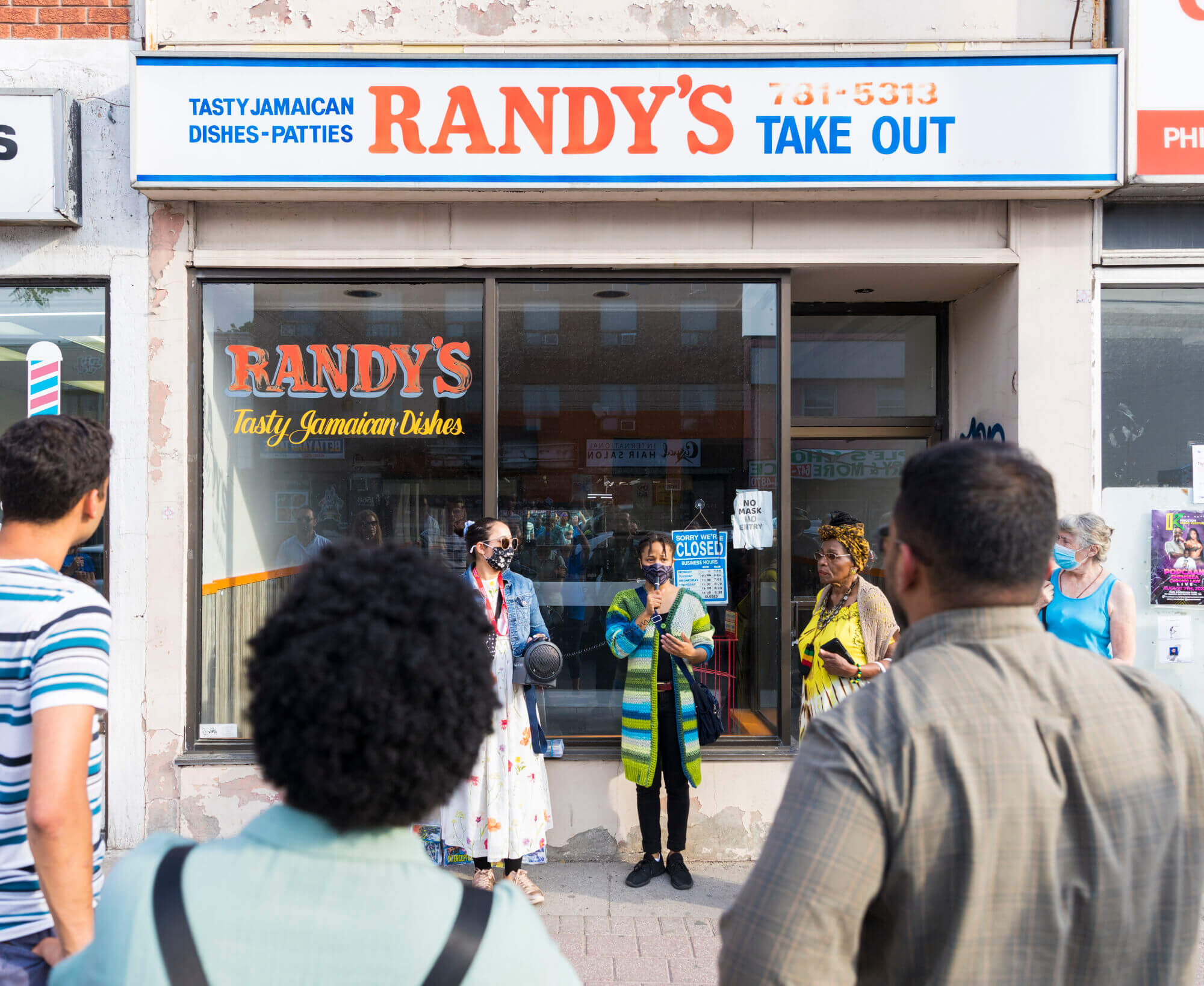 A woman is speaking to a group of people with a microphone and two people are standing beside her in front of a store with glass window panels at the storefront. The store sign above the windows reads 