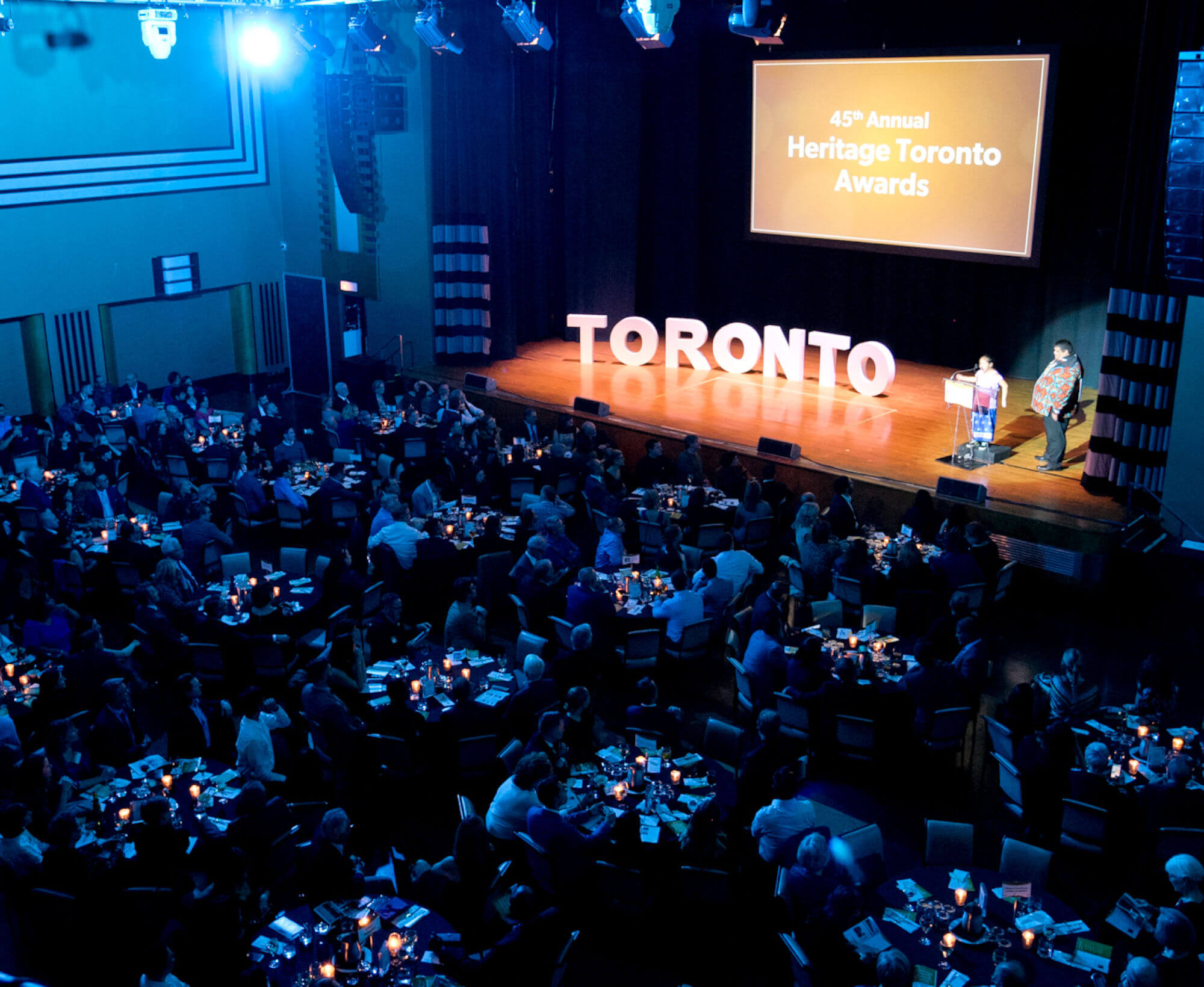 A photo taken from above looking down on a stage with a man and girl standing at the podium and large white letters spelling out Toronto to his left. Above and behind is a screen projection with the words 45th Annual Heritage Toronto Awards. On the floor of the auditorium are groups of people sitting at round tables.