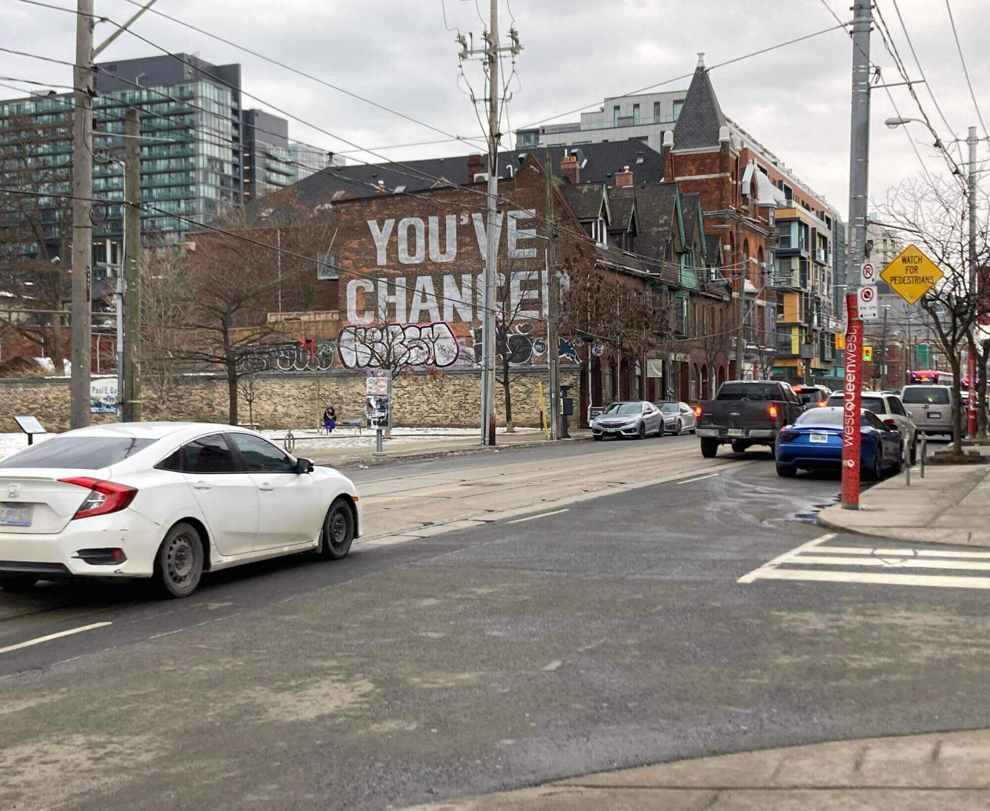 Behind some trees a brick wall has large white graffiti which reads 