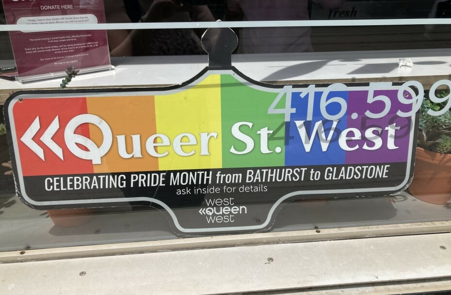 A sign shaped like an old-fashioned street sign with rainbow background sits on a counter behind a glass window. The sign reads “Queer St. West. Celebrating pride month from Bathurst to Gladstone. Ask inside for details. West Queen West.”